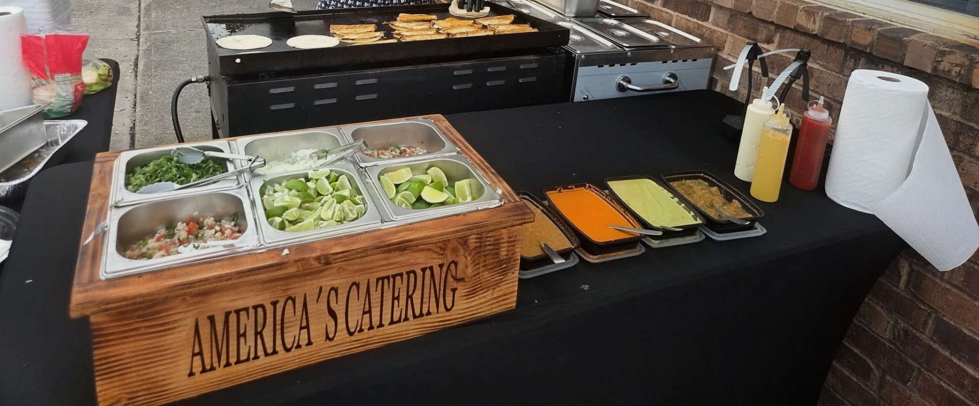 America's Catering taco bar setup with fresh salsas, limes, cilantro and onions beside the outdoor griddle